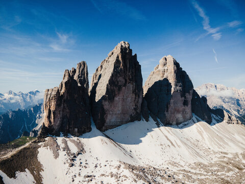 Aerial view of Tre Cime di Lavaredo, the iconic three peaks of the Dolomites with rugged rock faces, patches of snow, and clear blue sky in the background, Italy.