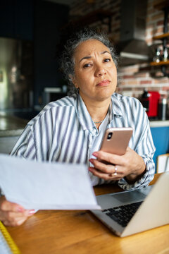 Concerned woman reviewing bills with smartphone at home kitchen