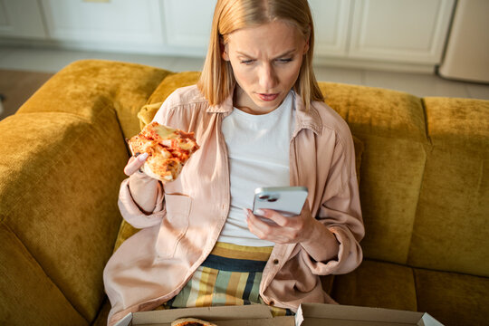 Young woman eating pizza while using smartphone on couch at home