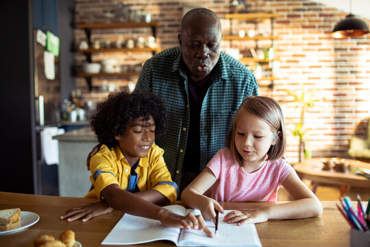 Older man helping children with homework in home kitchen