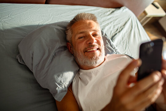 Smiling middle aged man lying in bed using smartphone at home