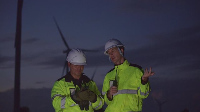 A professional Windmill engineer inspects and discusses operations at a wind turbine farm at dusk. Eco technology for electric, industry environment, sustainable technology wind power maintenance.