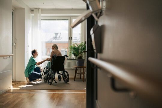 Happy female nurse talking to senior woman while crouching next to her wheelchair near window