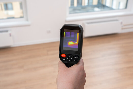 Hand holds a thermal camera. Screen shows color-coded heat patterns. Room has wooden flooring and windows. Professionals use it for energy audits. It highlights thermal imaging applications