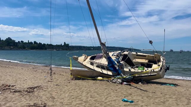 A wrecked sailboat lies abandoned on the sand at Kitsilano Beach, caution tape around its damaged hull, freighters anchored across the bay under a bright sky