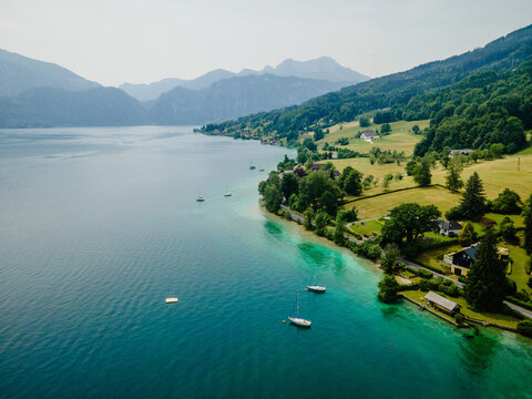 Aerial view of Attersee lake with sailboats on turquoise water, surrounded by green hills, meadows, and houses in Vocklabruck District, Upper Austria, Austria.