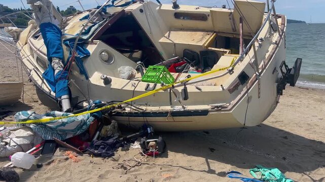 A wrecked sailboat heeled on the sand at Kitsilano Beach, cockpit exposed and belongings spilled across the beach under yellow caution tape, the bright bay beyond