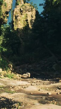 A dry riverbed shows scattered rocks and pebbles with tall rocky cliffs on one side. Sunlight casts shadows on the ground. Green vegetation grows along the edges of the scene.