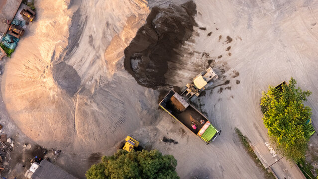 Aerial view of a construction site with a loader filling a dump truck with sand and gravel, surrounded by piles of earth and industrial equipment in Indonesia.