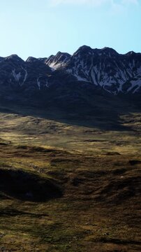During daytime, mountains tower over green hills in a remote location. The landscape shows the contrast between the rugged mountains and the flat terrain below.
