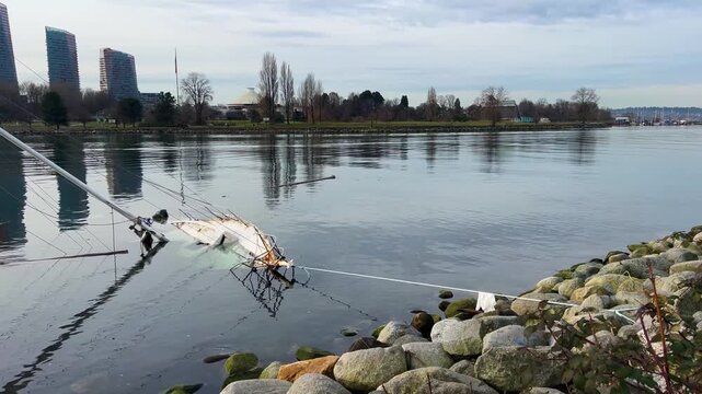 A capsized sailboat lies wrecked by a rock breakwater, the domed Space Centre, bare trees and downtown towers across calm winter water at English Bay in Vancouver.