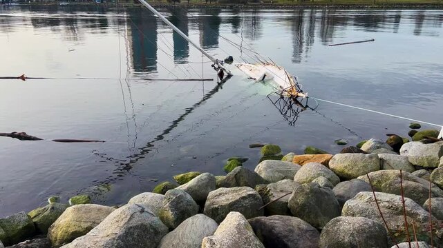 A capsized sailboat lies half-submerged beside a mossy rock breakwater, its fallen mast and rigging spread across calm reflective winter water at English Bay