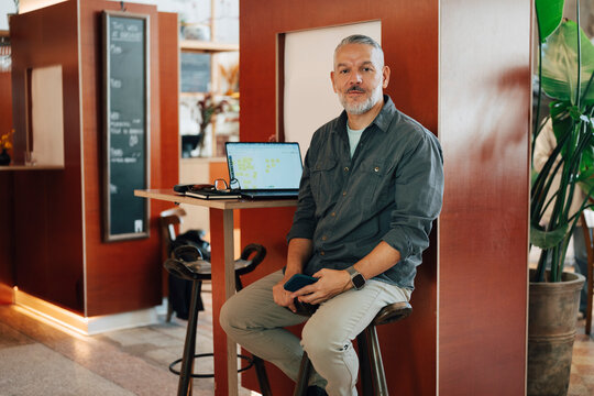 Portrait of mature male entrepreneur sitting on stool next to his working desk at shared workspace