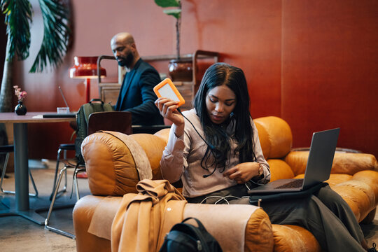 Female entrepreneur connecting external hard drive to her laptop while sitting on leather sofa at coworking space
