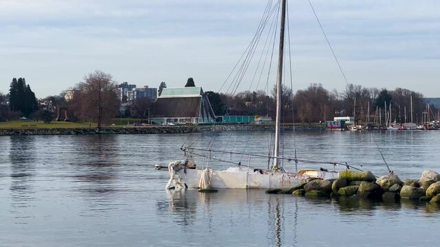 A wrecked sailboat lies grounded and broken on a rock breakwater, the green-roofed Vancouver Maritime Museum and Heritage Harbour across calm winter water