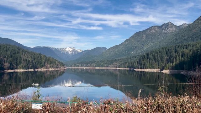 A flock of gulls glides over the mirror-calm Capilano Reservoir, snow-capped peaks and evergreen slopes reflected in the still water above Cleveland Dam in North Vancouver under a bright sky.