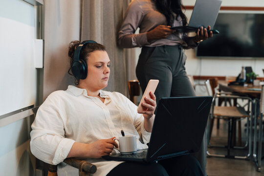 Female freelancer using her smart phone with laptop open on her lap at modern coworking space