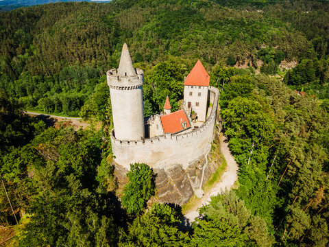 Aerial view of Kokorin Castle with its distinctive cylindrical tower and red-tiled roofs set atop a rocky outcrop amidst lush forest in Kokorin, Central Bohemian Region, Czechia.