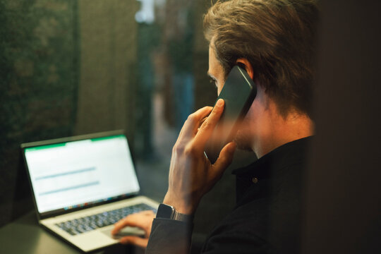 Businessman talking on smart phone while working on laptop inside glass cubicle