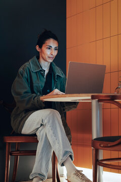 Full length of focused businesswoman working on laptop while sitting near tiled wall in illuminated coworking space loun