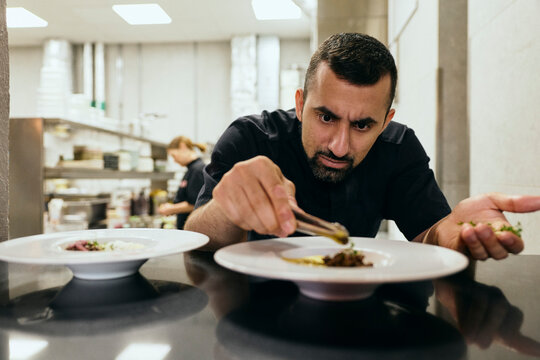 Focused male chef carefully placing microgreens on dish with tweezers in restaurant kitchen