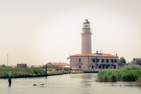 Scenic view of the historic Goro Lighthouse (Faro di Goro) located on the Isola dell'Amore in the Po River Delta, Italy, surrounded by calm water and coastal vegetation under a clear sky.