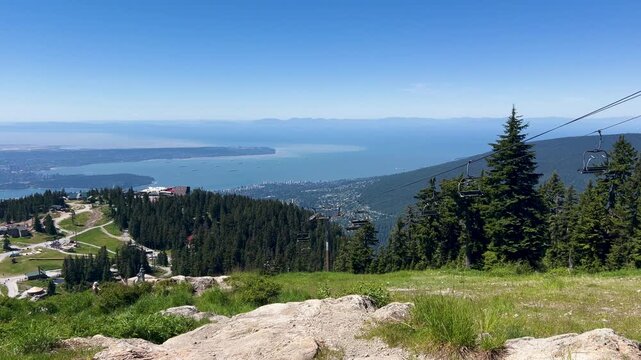 A grassy summit viewpoint at Grouse Mountain looks out over the city, English Bay and anchored freighters, a chairlift cable at the edge of frame on a summer day