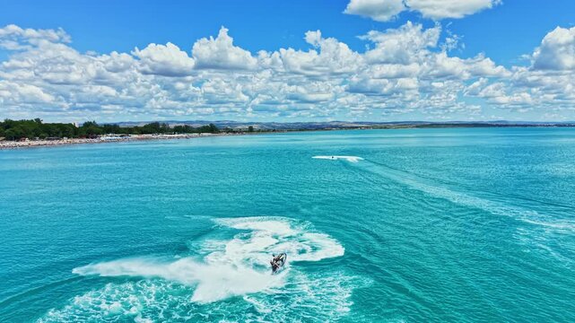 Boats create circular patterns in the blue water with waves while clouds float in the sky on a sunny day at the lake. The shore is visible in the background alongside green trees.