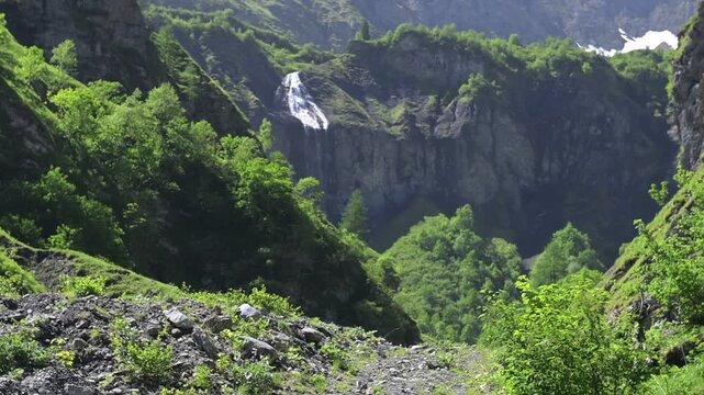 Scenic Swiss mountain gorge with distant waterfall, lush trees, and rocky slopes under bright summer sunlight.