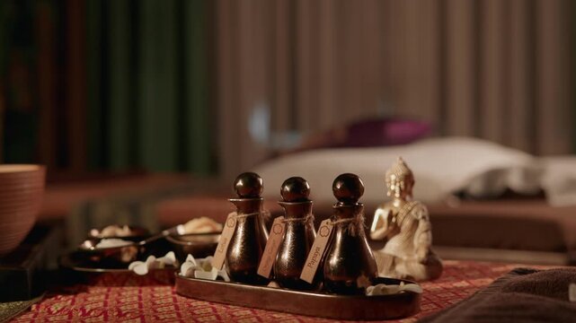Spa oils and buddha statue arranged on patterned fabric treatment table in tranquil indoor massage room with subtle bokeh background, warm light.