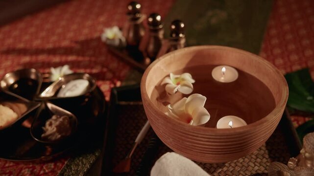 Slow motion close up of Thai relax wooden bowl with floating candles and plumeria flowers on spa tray in treatment room.