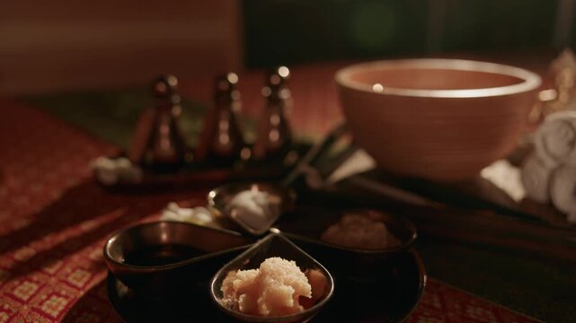Slow motion close up of spa bowls scrubs and oils arranged on patterned tablecloth in indoor massage room, warm light.