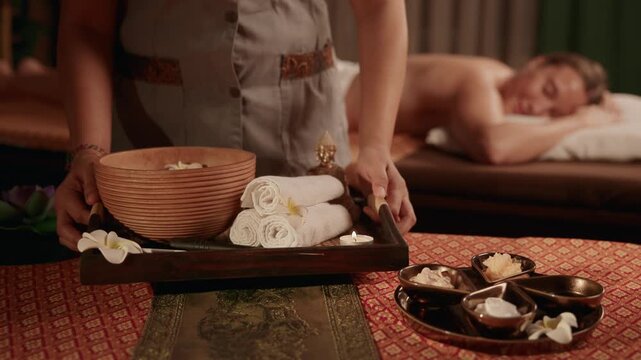 Slow motion spa therapist placing tray with towels and wooden bowl beside client on Thai massage table in candlelit treatment room.