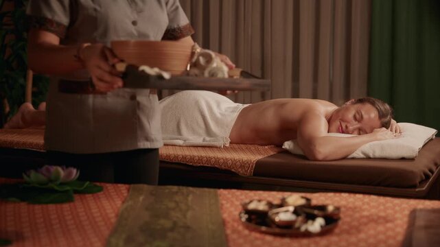 Tracking shot spa Thai therapist carrying aromatherapy bowl and rolled towels to woman lying on massage table in dim treatment room slow handheld movement.