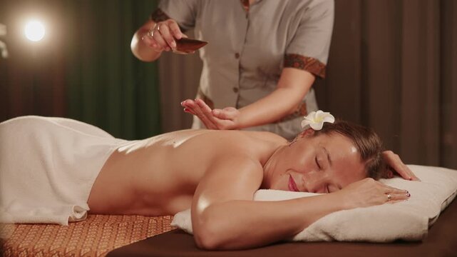 Slow motion Thai therapist pouring oil onto woman receiving back massage on spa treatment table in dim lit room.