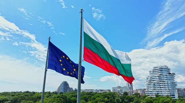 Flags of Bulgaria and the European Union fly on poles with a city in the background. The sky is blue with some clouds during the day. Buildings are visible below the flags.