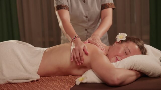 Happy woman receiving back massage on spa table in Thai treatment room with therapist hand movements.