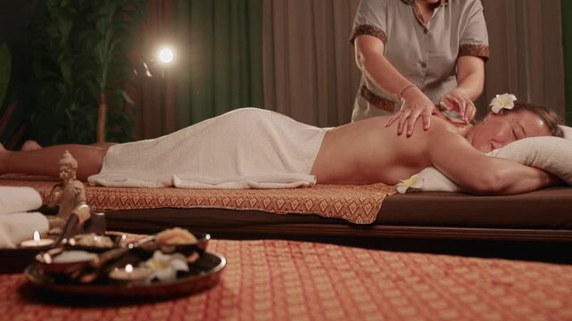 Slow motion Thai therapist massaging woman lying on aroma oil spa treatment table in dim candlelit massage room.