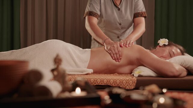 Woman receiving back massage on Thai spa treatment table in dim treatment room with candles and soft lighting, therapist hands moving along spine..