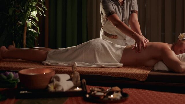 Thai therapist massaging woman on spa table in dimly lit treatment room with candles and plants, tracking shot.