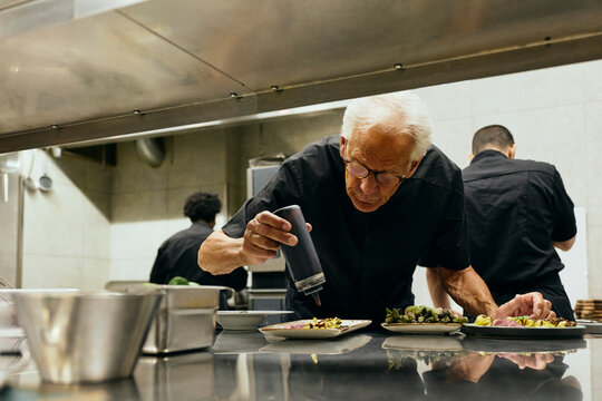 Senior male chef drizzling sauce over dish while working under steel kitchen hood