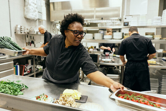 Busy female chef holding containers of fresh vegetables while working in commercial restaurant kitchen