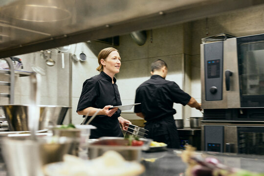Busy female chef holding plate and containers while working inside commercial kitchen