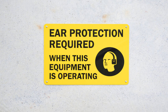 Yellow and black, ear protection required when this equipment is operating, text, sign on concrete wall