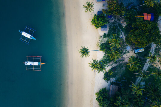 Aerial view of Port Barton featuring traditional outrigger boats on turquoise water near a sandy beach and palm trees in San Vicente, Mimaropa, Philippines.