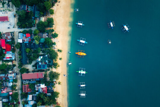 Aerial view of Port Barton with colorful outrigger boats moored along the sandy beach and lush tropical vegetation surrounding the coastal village in San Vicente, Mimaropa, Philippines.
