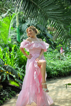Happy elegant woman in flying pink dress enjoying life with closed eyes in tropical garden. Cheerful emotional girl laughing in botanical greenhouse, care free concept.