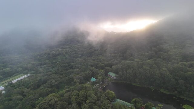 The sun rises over a forest with mist covering the trees near a quiet lake. Buildings are seen along the water's edge among the greenery.