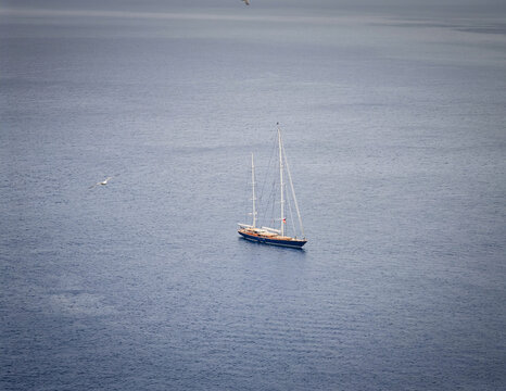 Aerial view of a serene sailboat floating on the open sea near the Capri island shore, featuring a blue and white wooden hull with metal masts, accompanied by a seagull.