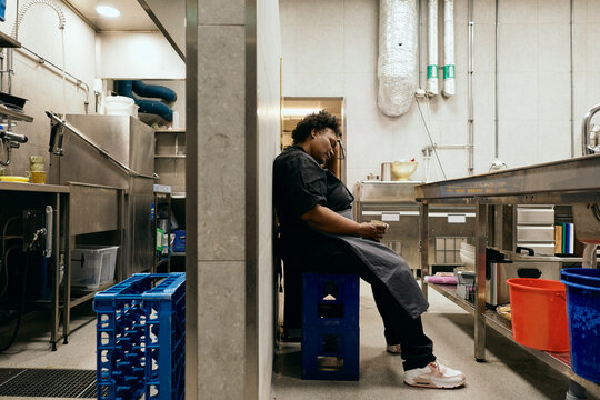Side view of exhausted female chef sitting on plastic crate during her shift in commercial kitchen